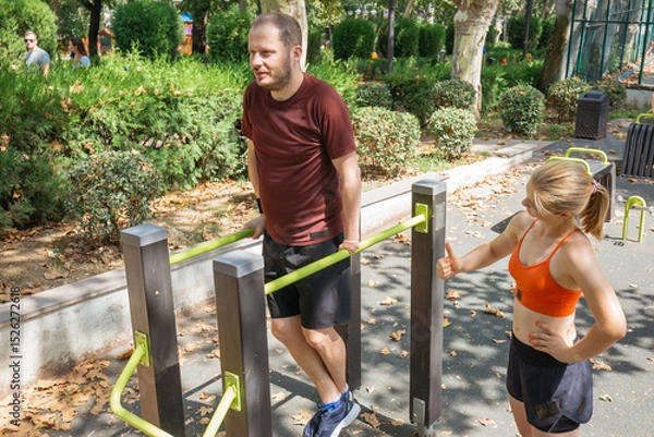 Obraz Man doing parallel bar dips outdoor in a park.  A woman assists him with his workout.  Calisthenics training and fitness in a public space.
