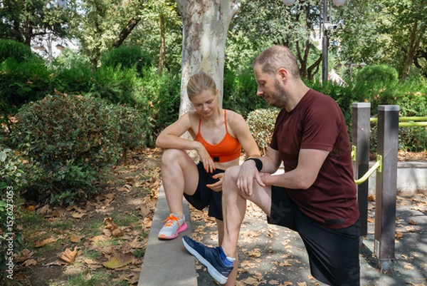 Obraz Young woman stretches her leg while her male trainer assists.  They're outdoors in a park, possibly in Europe, enjoying a sunny workout session. Perfect for fitness, sports, and health-related