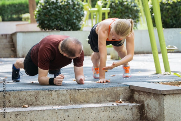 Obraz A fitness instructor assists a man performing a plank exercise outdoors. They are working out together in a park setting. The image depicts a healthy active lifestyle and personalized fitness