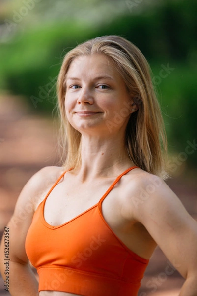 Obraz Portrait of a young blonde woman in an orange sports bra, smiling confidently.  She's outdoors with a blurred green background, suggesting a park or natural setting. The image evokes health, fitness,
