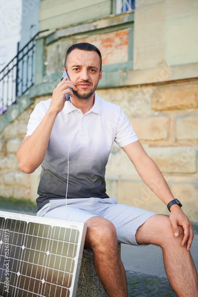 Fototapeta Handsome man using smartphone connected to photovoltaic solar panel. Integration of sustainable renewable energy into everyday life, demonstrating practical use of solar power for charging devices.