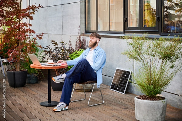 Fototapeta Happy bearded man works at round wooden table with laptop, notebook, and cup of coffee. Guy wears earbuds, surrounded by greenery and solar panel, highlighting modern, productive workspace.