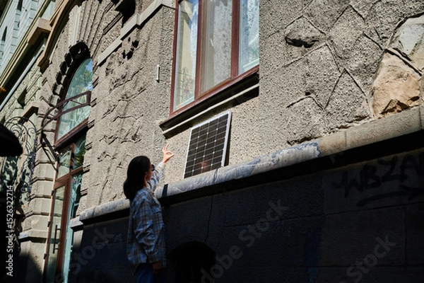Fototapeta Woman holding photovoltaic solar panel in front of historical building, showing Ok sign. Concept of integration of sustainable renewable energy sources into old architecture.