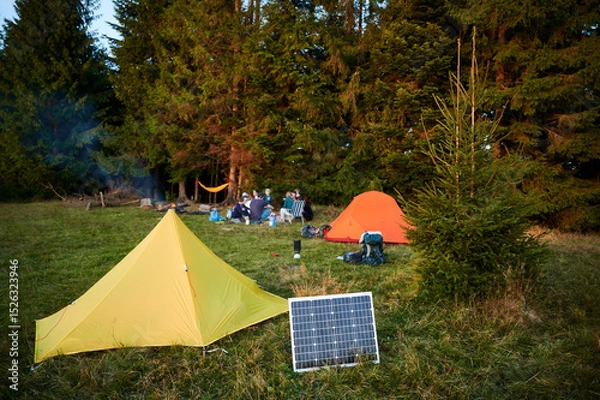 Fototapeta Solar panel in grassy field with tents in background highlights eco-friendly camping setup. Scene includes campers, emphasizing integration of renewable energy in outdoor activities.