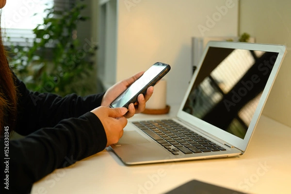 Fototapeta Close up shot of woman using smartphone at work desk with laptop