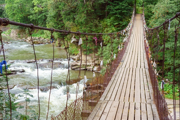 Fototapeta Wooden suspension bridge with colorful ribbons hanging from wires crosses fast mountain river, mountain creek in lush green coniferous Carpathian forest. Adventure, travel, hiking and local charm.