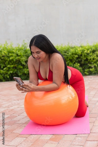 Fototapeta Latin woman using smartphone while doing pilates with an exercise ball outdoors