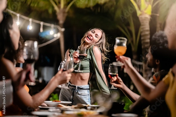 Fototapeta Group of diverse friends raising glasses in a joyful toast at a tropical outdoor party, celebrating together under palm trees and string lights