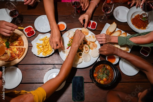 Fototapeta Close-up of friends sharing a delicious meal and drinks at a lively dinner party, featuring seafood, fries, and wine on a wooden table