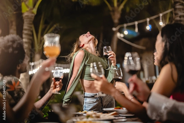 Fototapeta Group of diverse friends raising glasses in a joyful toast at a tropical outdoor party, celebrating together under palm trees and string lights