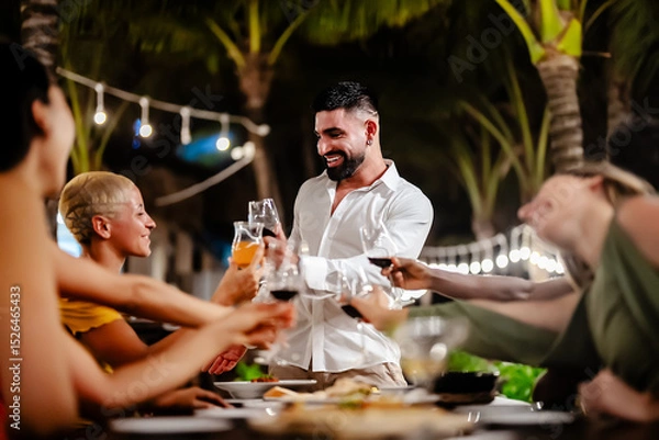 Fototapeta Group of diverse friends raising glasses in a joyful toast at a tropical outdoor party, celebrating together under palm trees and string lights