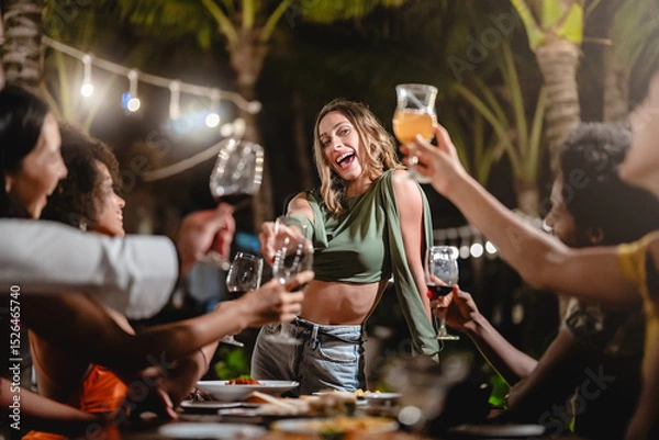Fototapeta Group of diverse friends raising glasses in a joyful toast at a tropical outdoor party, celebrating together under palm trees and string lights