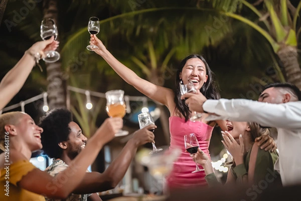 Fototapeta Group of diverse friends raising glasses in a joyful toast at a tropical outdoor party, celebrating together under palm trees and string lights