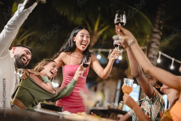Fototapeta Group of diverse friends raising glasses in a joyful toast at a tropical outdoor party, celebrating together under palm trees and string lights