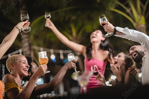 Fototapeta Group of diverse friends raising glasses in a joyful toast at a tropical outdoor party, celebrating together under palm trees and string lights