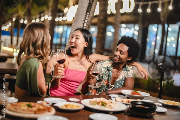 Fototapeta Group of happy friends sharing laughs and drinks at an outdoor dinner party under palm trees and string lights, enjoying food and festive vibes during a tropical night celebration