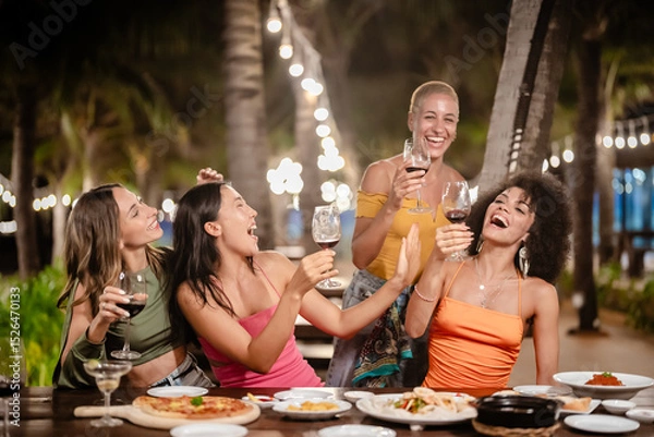Fototapeta Group of joyful women toasting with wine glasses at an outdoor tropical party at night, smiling and celebrating together under string lights surrounded by palm trees and a festive atmosphere
