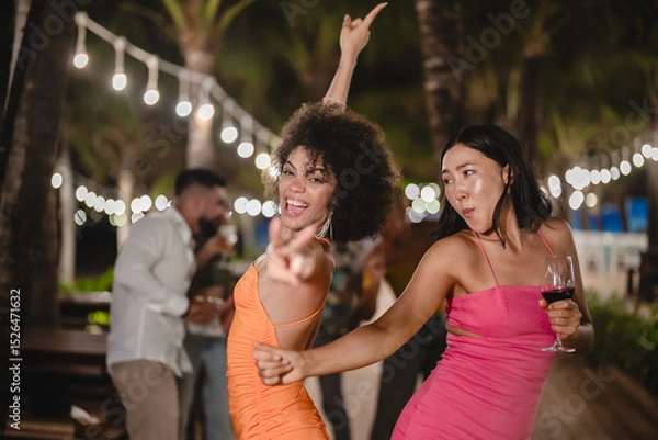 Fototapeta Two joyful women dancing with wine glasses during a fun tropical night party, surrounded by palm trees and string lights, celebrating friendship and enjoying the beach nightlife