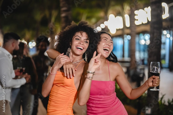 Fototapeta Two joyful women dancing with wine glasses during a fun tropical night party, surrounded by palm trees and string lights, celebrating friendship and enjoying the beach nightlife