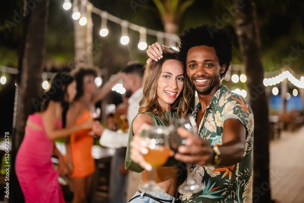 Fototapeta Smiling man and woman posing together while holding drinks at an outdoor night party, surrounded by friends dancing and celebrating under tropical palm trees and string lights