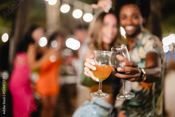 Fototapeta Smiling man and woman posing together while holding drinks at an outdoor night party, surrounded by friends dancing and celebrating under tropical palm trees and string lights
