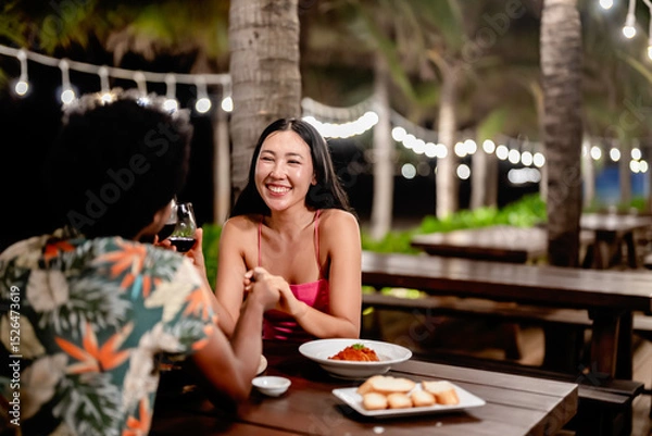 Fototapeta A couple toasting red wine over a romantic dinner at a tropical outdoor restaurant, featuring pizza, fries, and warm lighting for a cozy, intimate dining experience