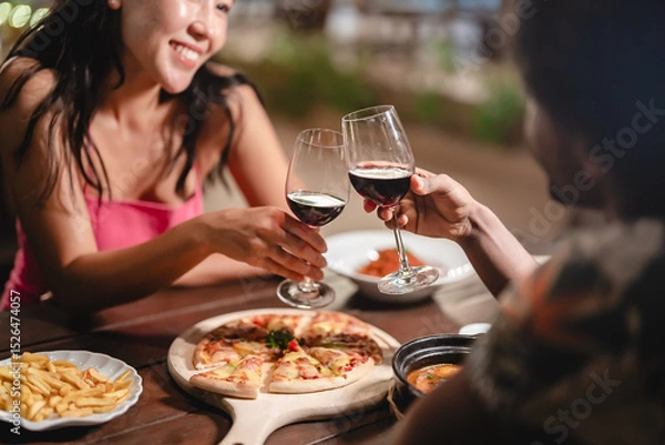 Fototapeta Close-up of a couple toasting red wine over a romantic dinner at a tropical outdoor restaurant, featuring pizza, fries, and warm lighting for a cozy, intimate dining experience