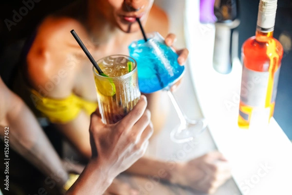 Fototapeta Close-up of two people enjoying colorful cocktails at a vibrant bar, featuring a blue tropical drink and a highball with lime, highlighting nightlife and summer party vibes