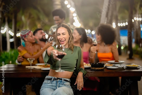 Fototapeta Joyful woman raising her glass at a festive outdoor dinner party with a diverse group of friends, celebrating and laughing under string lights in a tropical evening setting