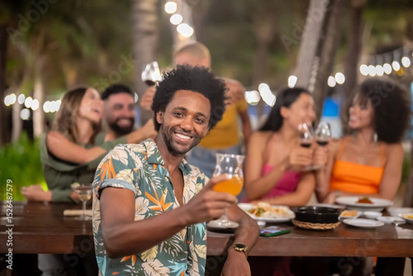 Fototapeta Smiling man raising a glass at an outdoor dinner party with friends, celebrating and enjoying food and drinks at night under string lights in a tropical setting