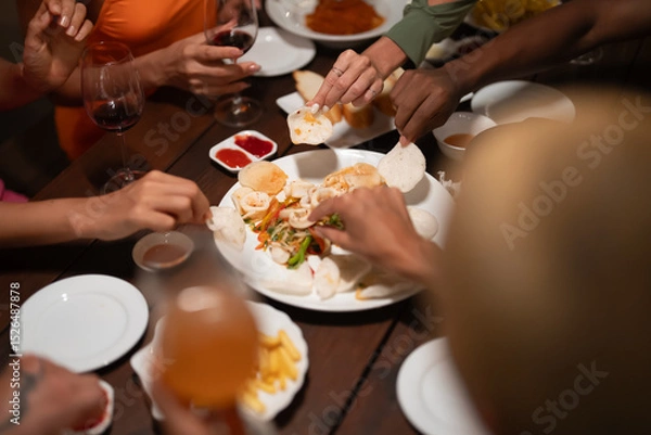 Fototapeta Close-up of friends sharing a delicious meal and drinks at a lively dinner party, featuring seafood, fries, and wine on a wooden table