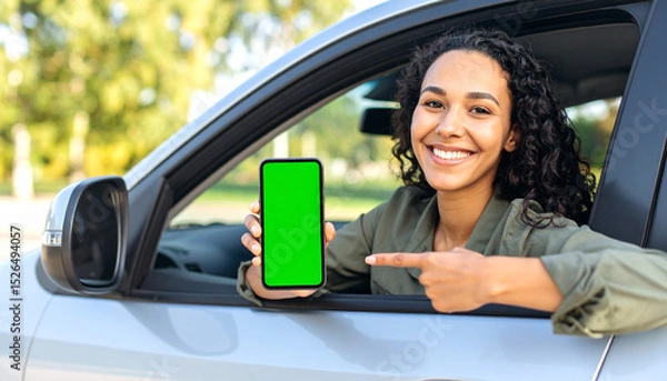 Fototapeta Woman in driver’s seat pointing at smartphone with green screen, showing device to camera, car interior, mobile app mockup concept

