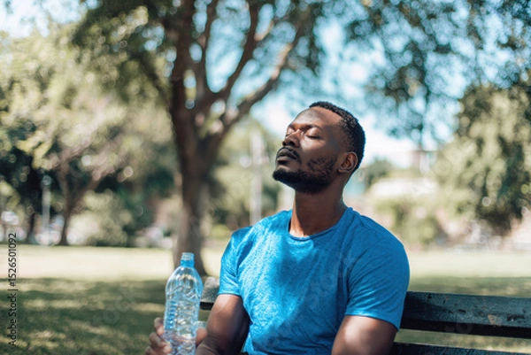 Fototapeta A man in his thirties relaxes on a park bench, enjoying a moment of peace under the sun while holding a bottle of water. He is coping with the intense summer heatwave