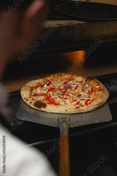 Fototapeta A close-up view of a pizza being removed from a stone oven using a classic wooden-handled peel. The perfectly puffed and charred crust, along with toppings like mushrooms, red bell peppers, and ham