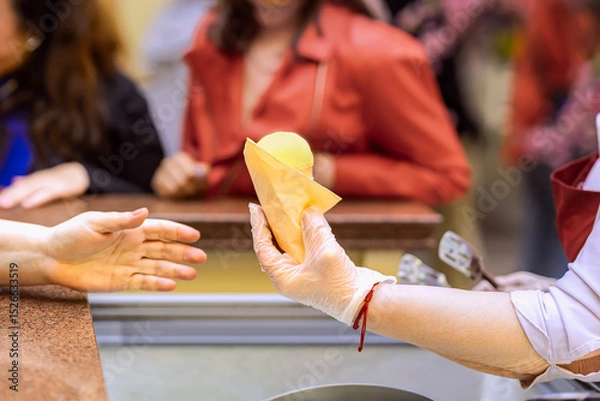 Fototapeta Ice cream seller giving ice cream cone to customer. Vendor in glove handing yellow ice cream cone to customer across counter. Concept of ice cream, food service, organic summer refreshment