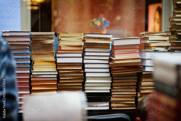 Fototapeta Tall piles of various books stacked together in indoor store setting. Tower of Knowledge, stacked wall of books. Concept of books, literature abundance, choice overload and printed knowledge