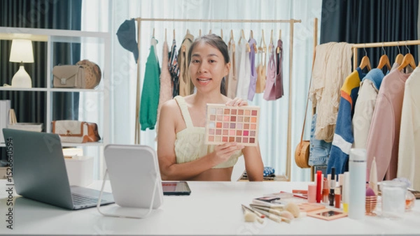 Fototapeta Young beautiful Asia lady blogger looking at camera recording swatch apply eyeshadow palette on skin show review to follower product vlog video live streaming at home. Business online media concept.