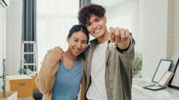 Fototapeta Young happy Asian sweet couple hugging each other smiling looking at camera show proud show off new house keychain in living room. Husband wife move new home, Apartment Rent And Ownership Concept.