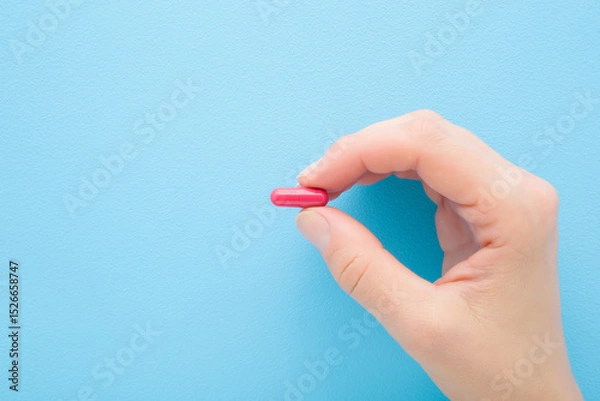 Fototapeta Young adult woman hand fingers holding and showing red pill for improve of iron level in blood on light blue table background. Pastel color. Closeup. Top down view.