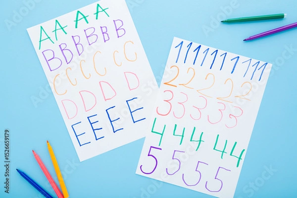 Fototapeta Colorful markers and written different english letters and numbers on white paper sheets on light blue table background. Pastel color. Closeup. School exercises for little child. Top down view.