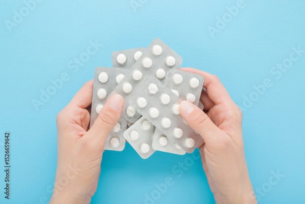 Fototapeta Young adult woman hands holding and showing blister packs with white pills on light blue table background. Pastel color. Closeup. Receiving vitamins or medication. Point of view shot. Top down view.