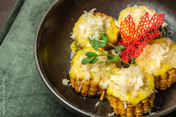Obraz Grilled corn medallions topped with melted cheese, shaved parmesan, microgreens, and decorative garnish served in a dark ceramic bowl.