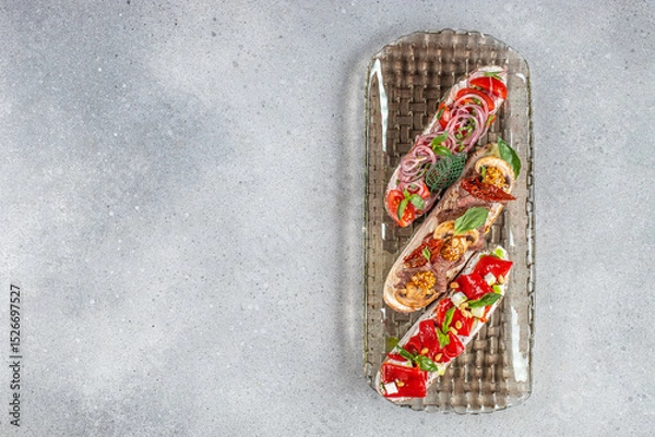 Obraz Gourmet bruschetta trio with fresh toppings including vegetables, herbs, meat, and cheese served on a textured glass platter over a stone surface