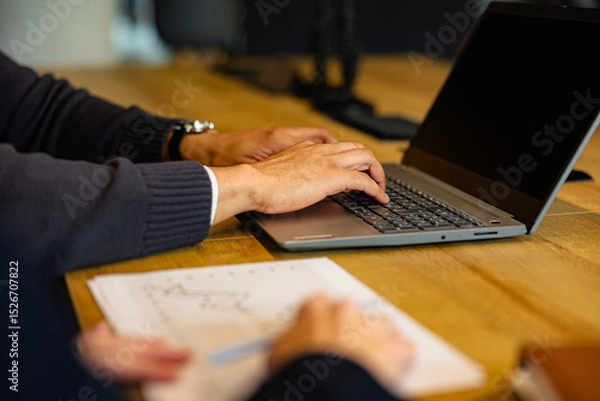 Fototapeta Businessman working on a laptop, analyzing financial data while a colleague diligently taking notes during a strategic office meeting