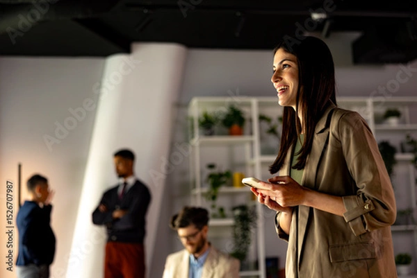 Fototapeta Businesswoman using mobile phone and smiling while her colleagues are standing in the background in a modern office