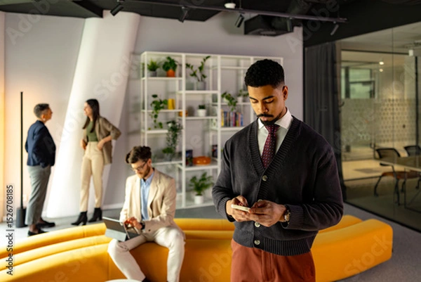 Fototapeta Young businessman texting on his phone while his colleagues work in the background in a modern office