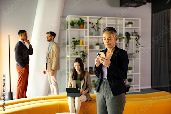 Fototapeta Mature businesswoman using mobile phone in modern office environment, with colleagues discussing and working in the background
