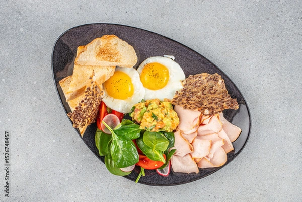 Obraz Healthy breakfast plate with sunny side eggs, avocado mash, greens, turkey slices, and toast.