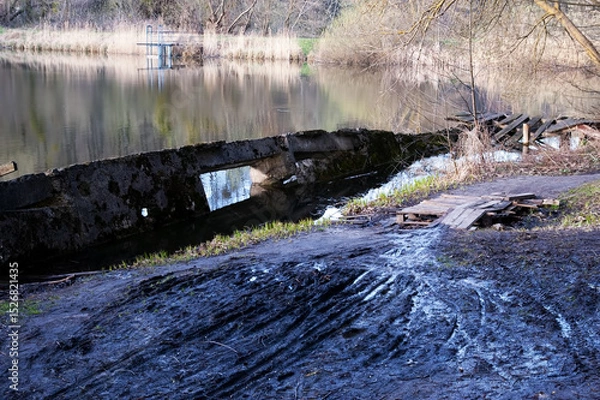Fototapeta Lake with sinking concrete wall. Muddy ground with narrow tire tracks. Small stream crossed by wooden pallet bridge. Dry reeds on far shore, path continues beyond bridge.