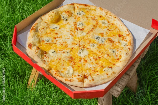 Fototapeta Cheese pizza in a red box on a stool on the grass. Food delivery, pizza.